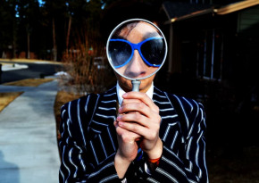 Man in blue suit with black stripes and blue sunglasses looking at the camera through a magnifying glass